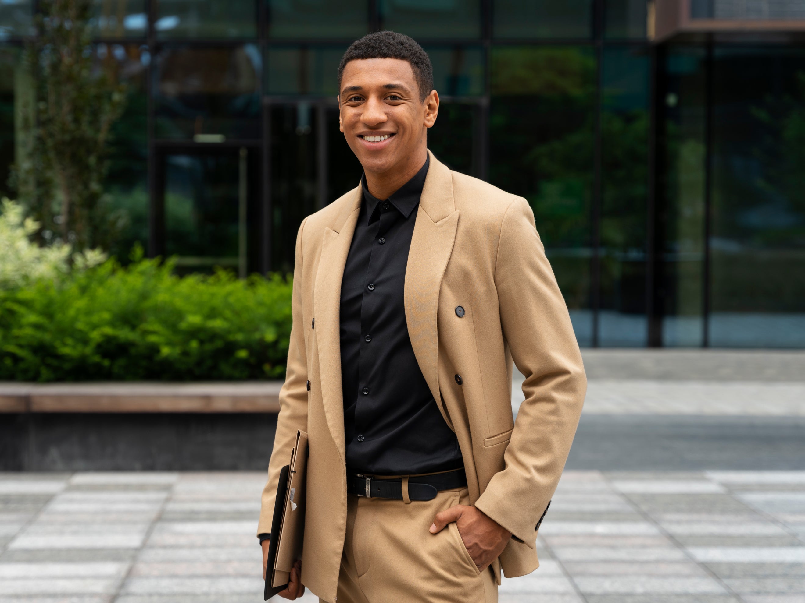 Man in a beige blazer and black shirt standing outdoors with a blurred background